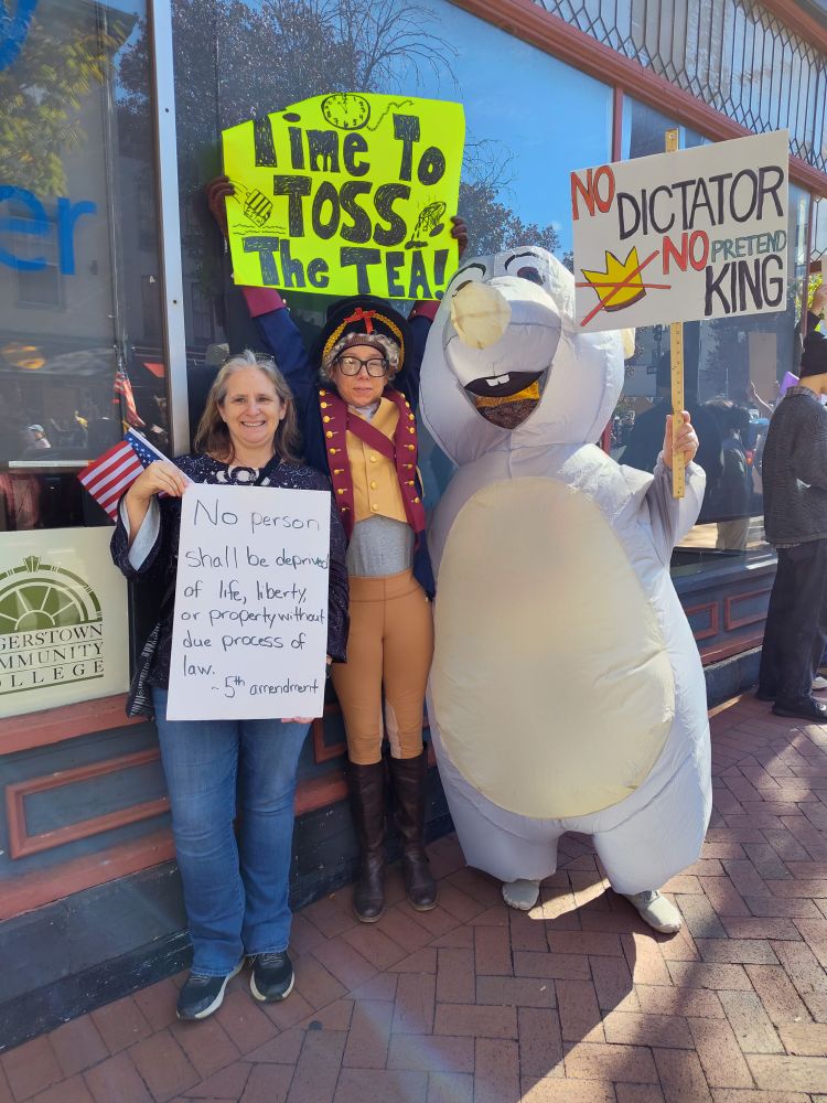 Three women hold protest signs at the No Kings protest, one in an inflatable mouse costume and on dressed like George Washington.