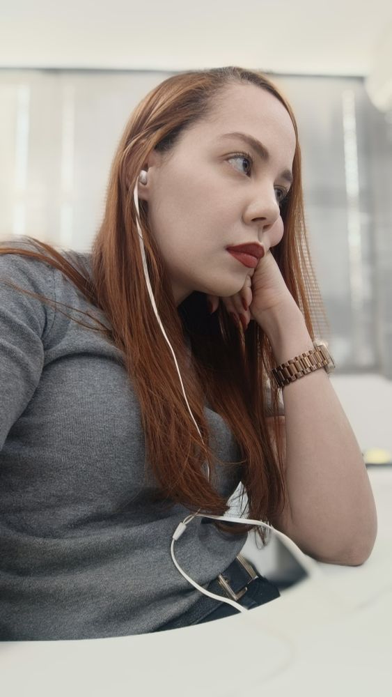 Mulher de perfil. Ela tem cabelos ruivos e está usando uma camiseta de tricô cinza e um relógio no braço