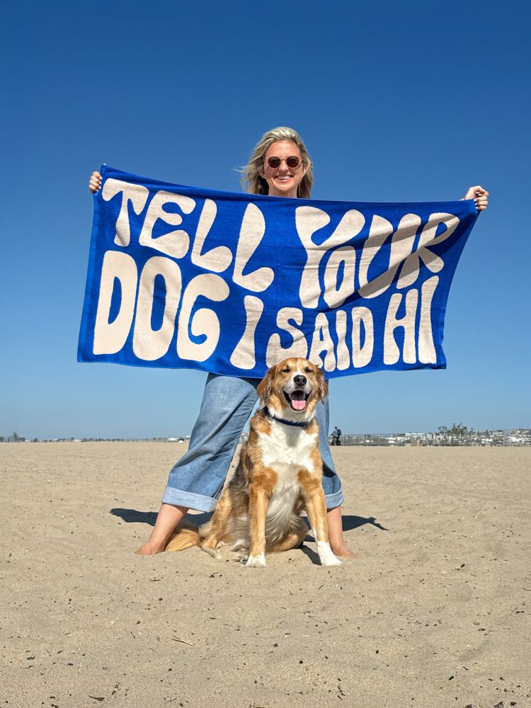a blond woman wearing jeans stands on the beach in a wide stance. she holds a blue 'tell your dog i said hi' towel out in front of her. a brown and white dog sits between her legs and smiles at the camera.