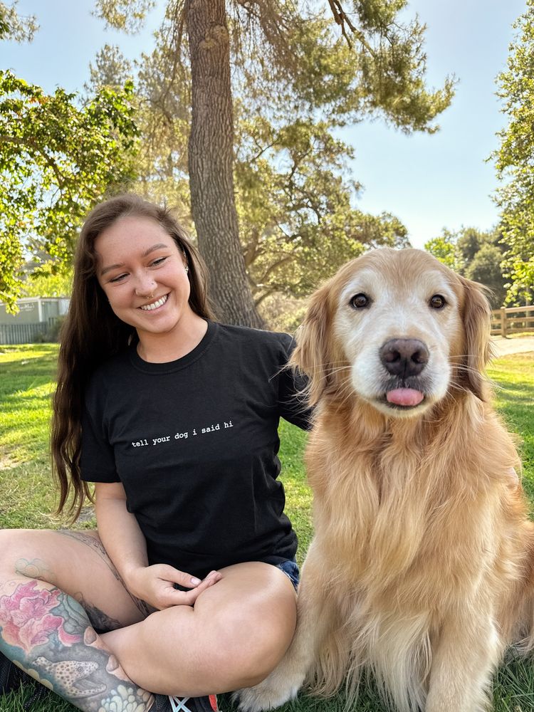 the same golden, Fresno, is now sitting up facing the camera. he is next to the human wearing a black shirt that says “tell your dog i said hi” across the front. Fresno’s tongue is slightly poking out of his mouth. 
