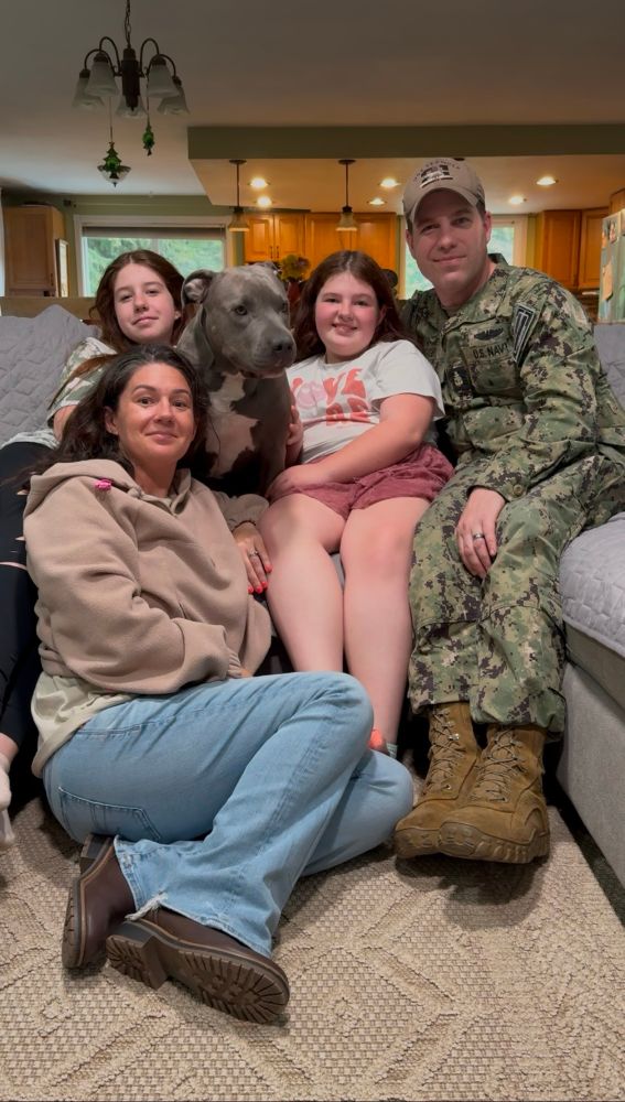 Juno poses on a couch with her entire family. there are two adults and two children. all of them are smiling while Juno is a little distracted by something just off camera.