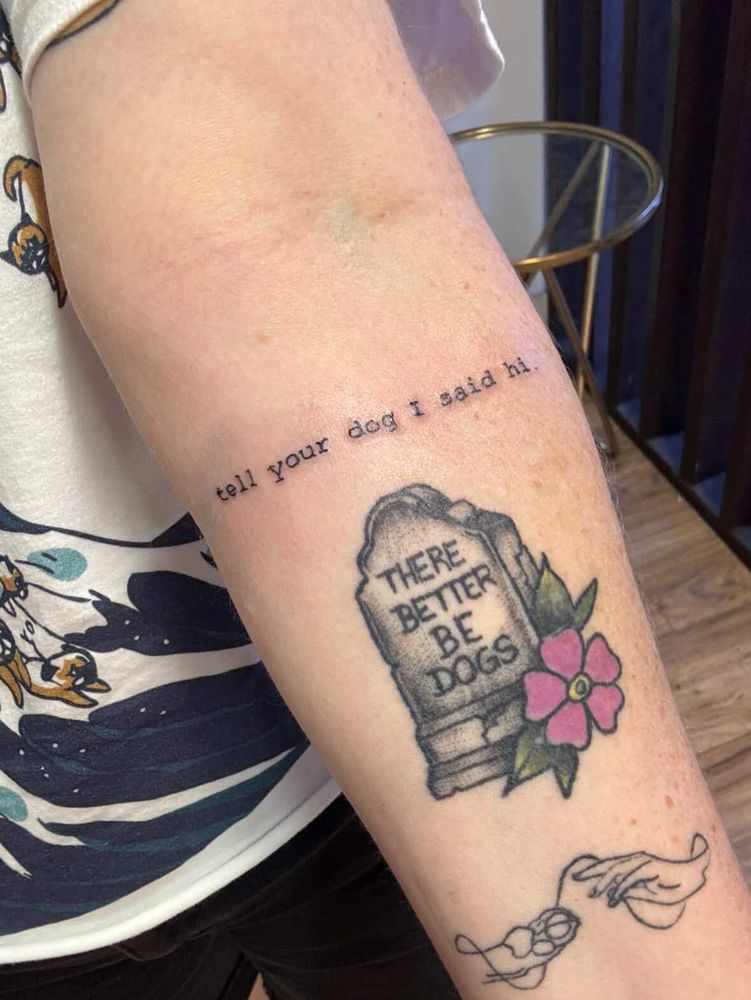 a person has a 'tell your dog i said hi' tattoo on their forearm just above another tattoo of a headstone that reads, 