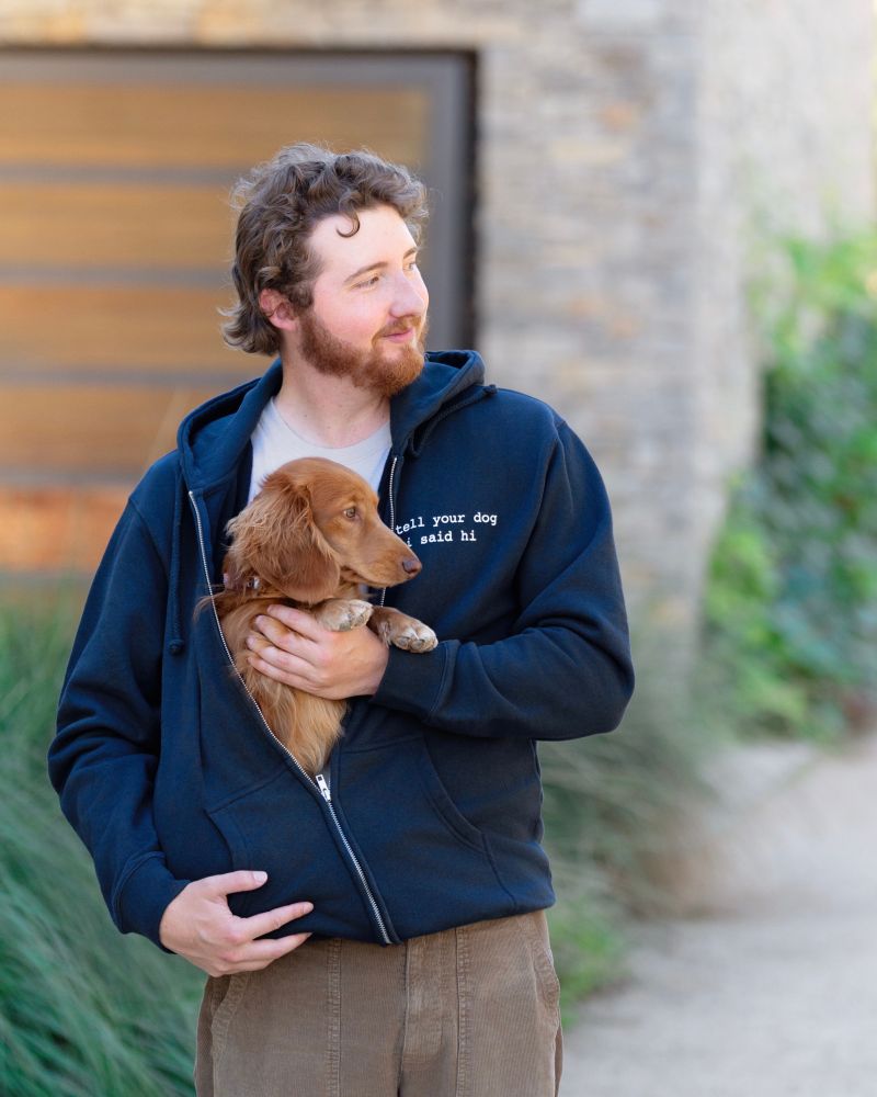 a man stands outside in a dark blue zip hoodie that reads "tell your dog i said hi". the light brown long hair mini-dachshund is partially zipped inside the hoodie. both are looking off camera in the same direction. 