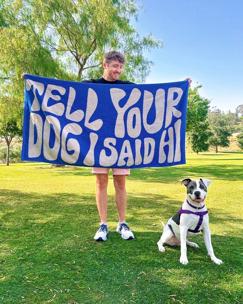 Matt, the founder of WeRateDogs, stands in a park surrounded by vibrant green grass and trees. he's holding a blue beach towel with wavy font that reads, 