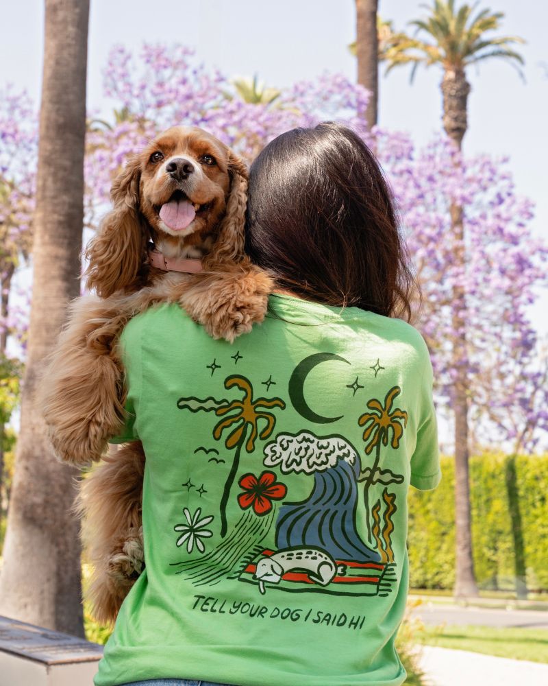 a tan cocker spaniel is being held by their human like a toddler over their shoulder. the pup smiles toward the camera. the back of the green 'tell your dog i said hi' shirt displays a sleeping dog in a summery scene featuring a big wave and moon.