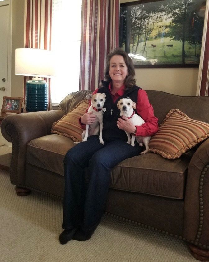 Macy poses for a family photo with her new mom and sister, Peaches. they're on a brown couch in their living room. both pups are looking at the camera while their mom holds them on either side of her and smiles.