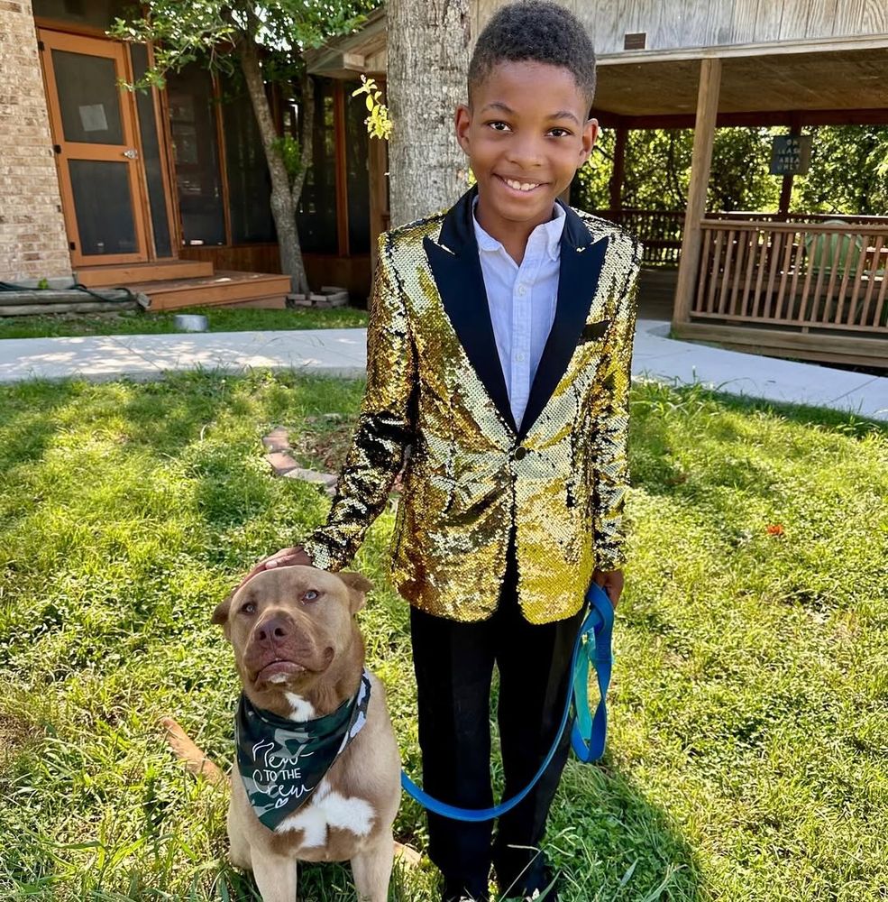 a photo of a kid dressed in a snazzy suit standing outside next to his pittie companion. the two look ecstatic to be in each others company 