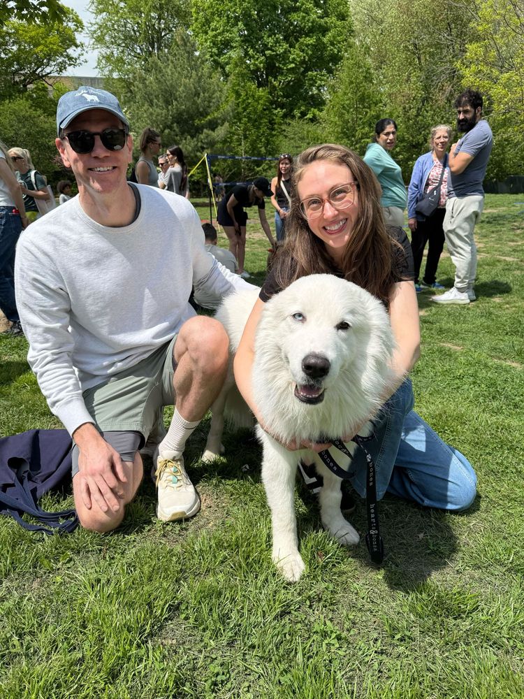 crocker is outside touching grass and smiling at the camera while being held by two smiling humans. 