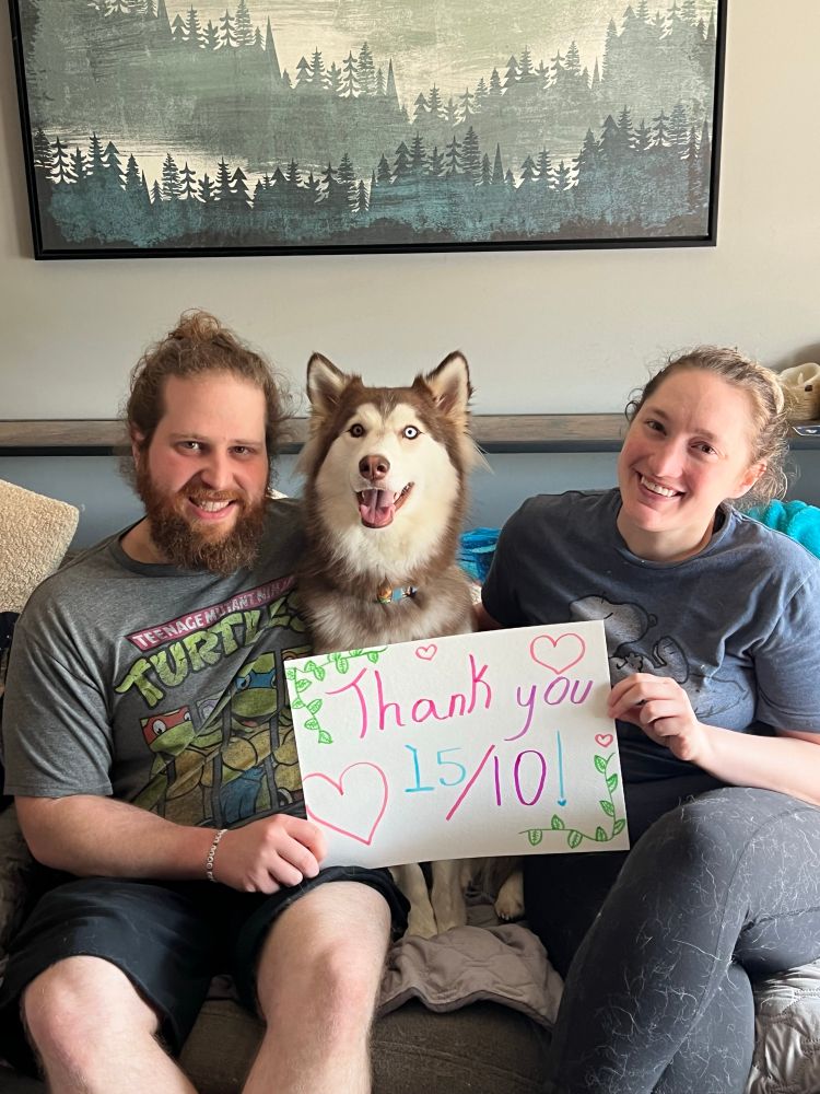 shelby sits on a cream couch surrounded by her two humans. they hold up a sign that says "that you 15/10." shelby is smiling like it's a class photo. 
