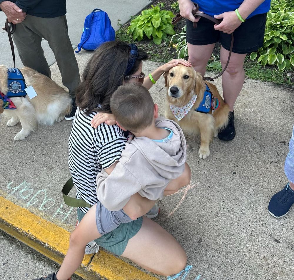 a person holding a child on their hip crouches down on the sidewalk to pet a golden retriever on the head.