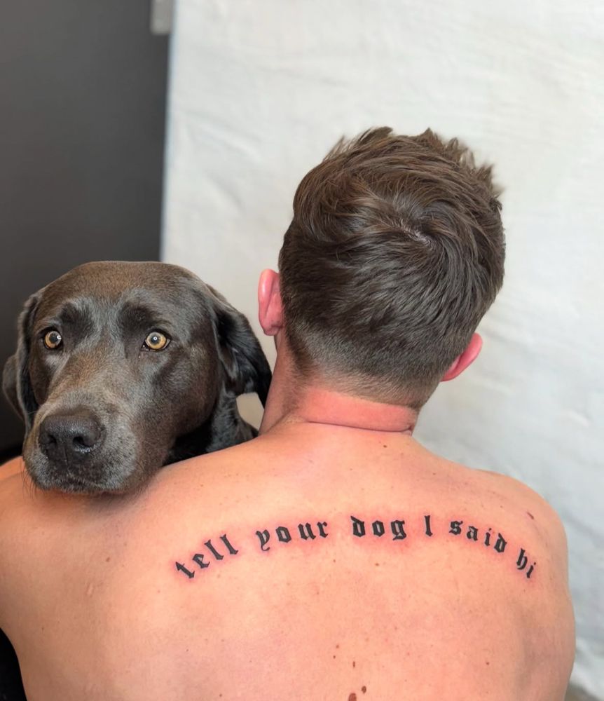 a man faces away from the camera while a gray lab peeks over his shoulder. the man has a fresh tattoo across his shoulder blades that reads 'tell your dog i said hi' in an old english font.