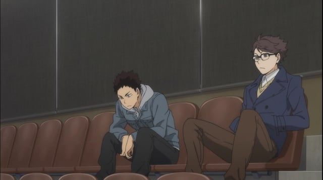 Oikawa and Iwaizumi from the anime Haikyuu!!, watching a volleyball match. Oikawa is wearing a dark blue double-breasted jacket over a white shirt and beige vest, brown trousers, and half-rim glasses. Iwaizumi is wearing a grey blue jacket, possibly denim, over a light grey hoodie and black trousers. Their height being above average is indicates by how high their knees are as they sit in the arena seats. They are seated with the seat between them deliberately left empty.