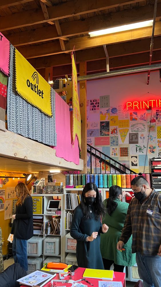 A colorful indoor studio space with walls covered with colorful paper and neon lights. There are people crafting an image for printmaking. 