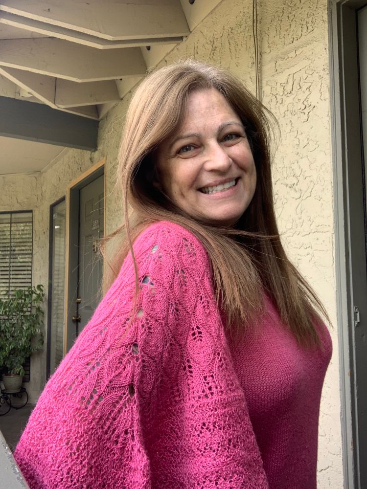Smiling woman wearing a knit she just completed, a bright pink sweater with an intricate lace bell sleeve. 