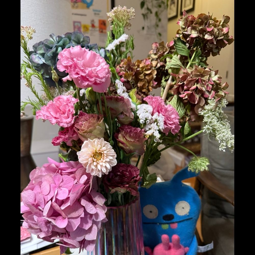 A beautiful flower arrangement of various pink colored flowers from dark to light to bright pink, in a pink fluted vase with a blue and a pink felt ugly doll standing beside the vase. 