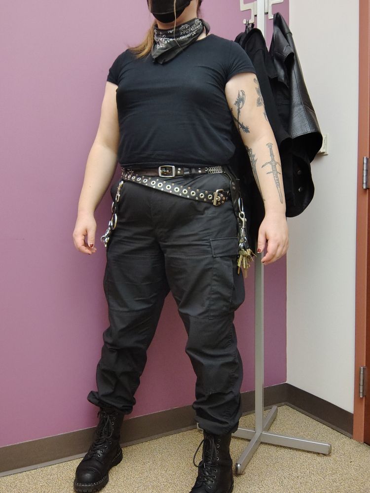 A picture of Jenn standing in front of a coat rack, dressed in a practical all-black outfit.