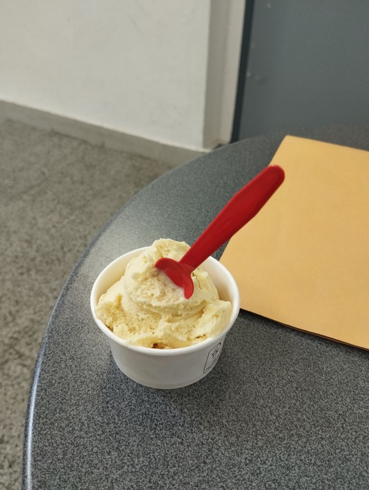 Auf einem grauen Tisch in einem Flur liegt ein gelber Hefter und ein kleiner Pappbecher mit einer Kugel Vanilleeis und einem kleinen roten Löffel. 