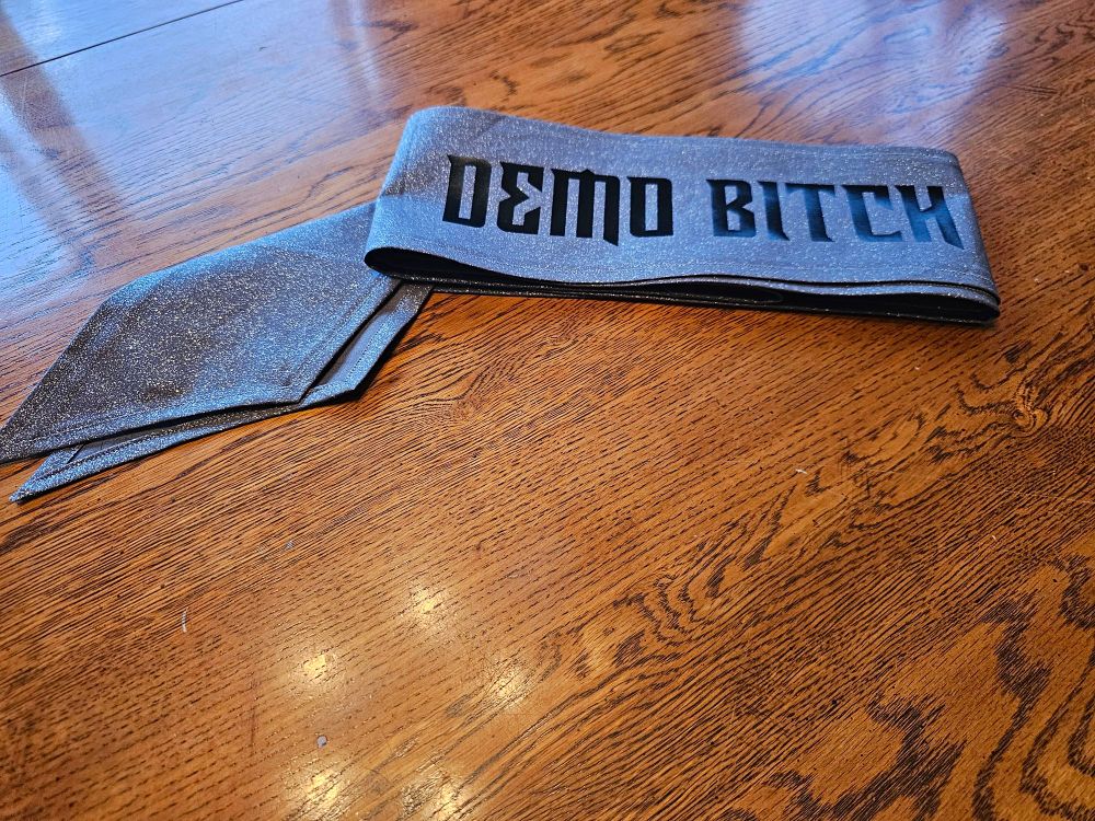 Picture of a glittery grey sash with the words "DEMO BITCH" printed on it.