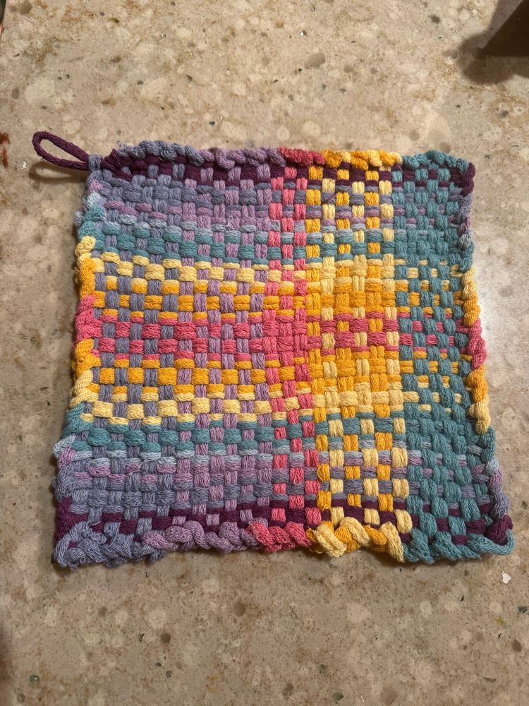 A handmade potholder of purple, violet, periwinkle, aqua, yellow, light orange, and carnation pink. 