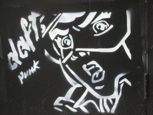 Photograph of street art. White spray paint on black background, on the left are the stylized words 'daft punk', the right side of the photo is dominated by the spray-painted image of a woman's face with a mask maybe...it's hard to say.