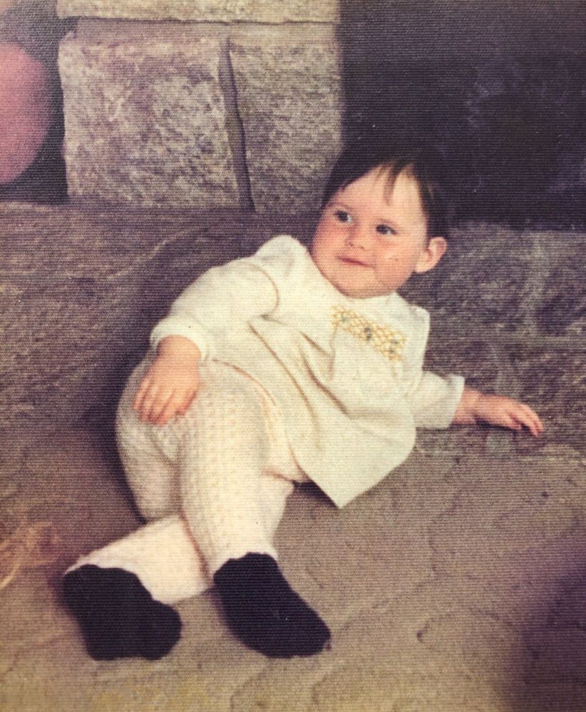 Photo of Elizabeth at 1 yrs old. The baby is dressed in a hand knit white outfit and she's lounging in front of a fire place.