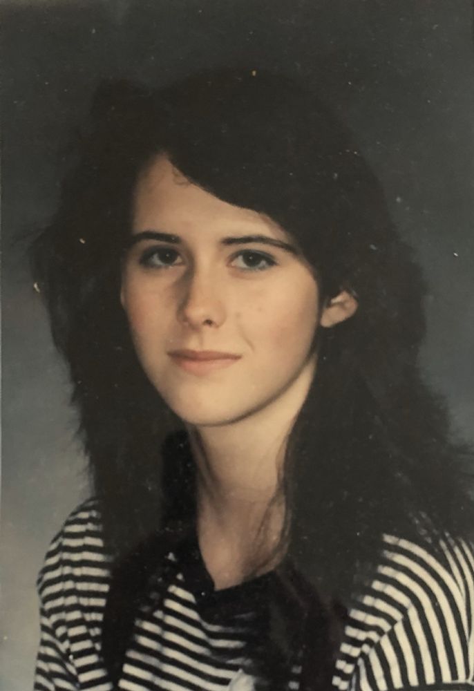 Photo of Elizabeth at 15 yrs old. She is wearing her hair long and in layers, as was the style at the time. It's a school photo, so she refuses to smile to reveal her braces. She's wearing a black and white striped top.