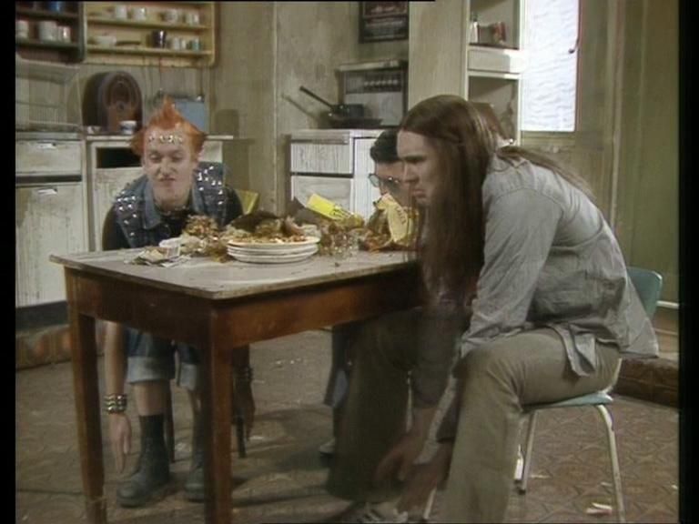 A still from The Young Ones. Three men sit around a table in a dirty kitchen, all thrusting their hands towards the floor. This being their response to their flatmate saying "hands up who like me."