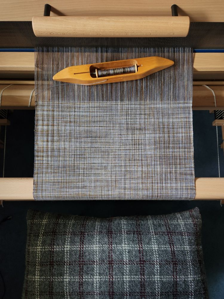 Overhead view of a weave in progress on a floor loom. The weaving bench has a cushion covered with handwoven wool cloth 