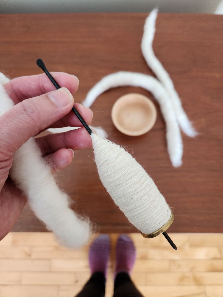 A hand holding a tahkli spindle. There is freshly spun natural white cotton on the spindle.