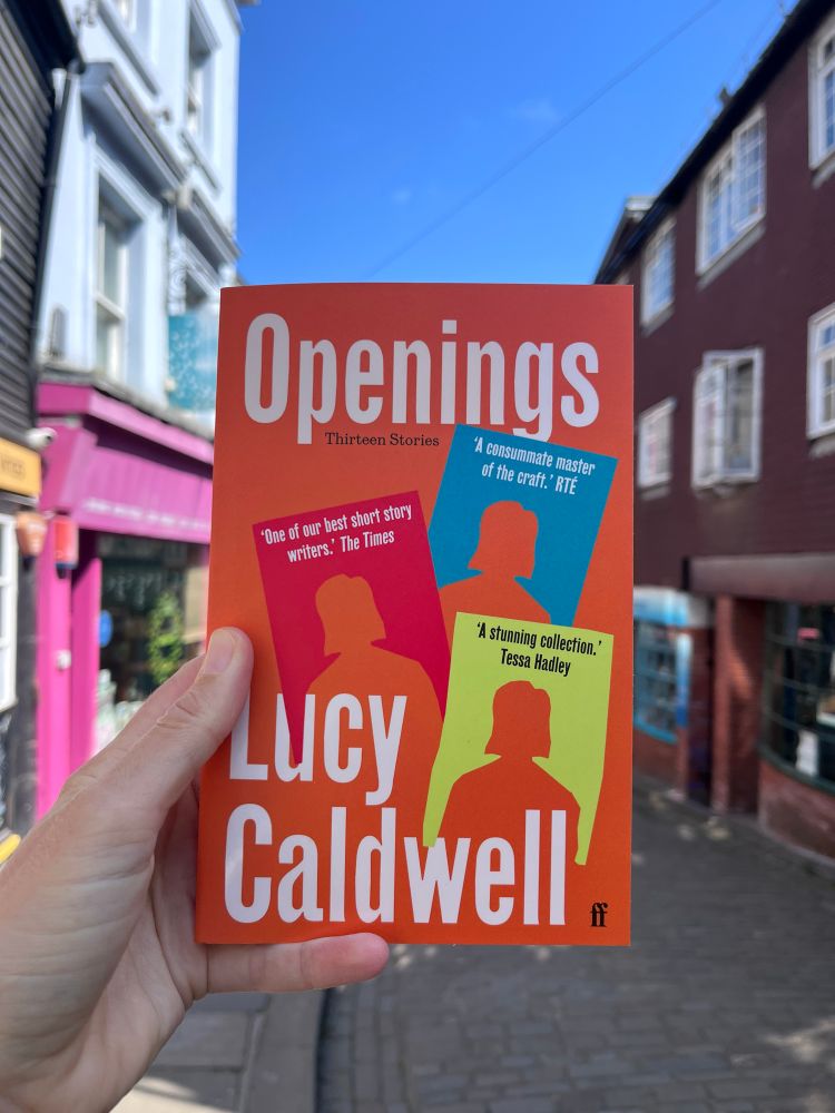 The brightly-coloured paperback of Openings held up against a colourful street and blue sky, all that’s missing is an aperol spritz and a sunlounger.