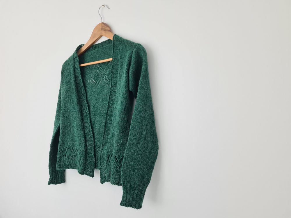 Green handknitted cardigan with a lace pattern at shoulders and hem. Shown from the front. 