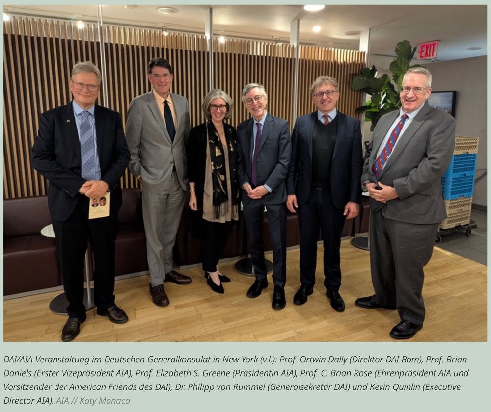 Sechs Personen stehen nebeneinander in einem modern eingerichteten Raum mit Holzwand und Pflanzen. Sie tragen formelle Kleidung, lächeln in die Kamera und posieren für ein Gruppenfoto. Die Bildunterschrift gibt Auskunft um wen es sich handelt: Prof. Ortwin Dally (Direktor DAI Rom), Prof. Brian Daniels (Erster Vizepräsident AIA), Prof. Elizabeth S. Greene (Präsidentin AIA), Prof. C. Brian Rose (Ehrenpräsident AIA und Vorsitzender der American Friends des DAI), Dr. Philipp von Rummel (Generalsekretär DAI) und Kevin Quinlin (Executive
Director AIA).