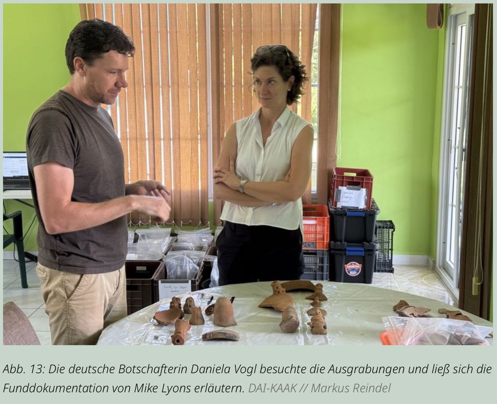 Die deutsche Botschafterin Daniela Vogl informiert sich bei einem Besuch der Ausgrabungen über archäologische Funde. Mike Lyons erläutert die Funddokumentation anhand ausgelegter Keramikfragmente.

Ein Mann (Mike Lyons) steht links an einem Tisch und spricht gestikulierend mit einer Frau (Daniela Vogl), die rechts im Bild mit verschränkten Armen zuhört. Auf dem Tisch liegen zahlreiche archäologische Fundstücke aus Ton, darunter Fragmente von Gefäßen und kleine Figuren. Im Hintergrund stehen beschriftete Kisten und Plastikboxen mit weiteren Fundmaterialien in einem Raum mit grünen Wänden und bodenlangen Jalousien.