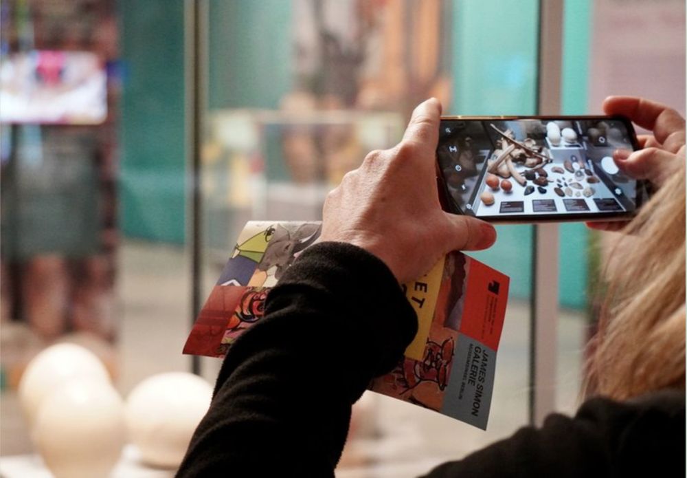 Foto, auf dem ausschnittsweise die Hände einer Person zu sehen sind, die ein Mobiltelefon halten, auf dessen Display die gerade damit fotografierte Ausstellungsvitrine zu erkennen ist.