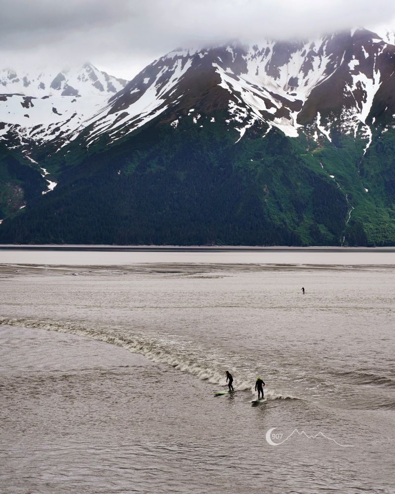 A photograph of two surfers riding the bore tide. The water is colored brown because of the strong current churning up the sediment. In the background is a green mountain with snow capped peaks.