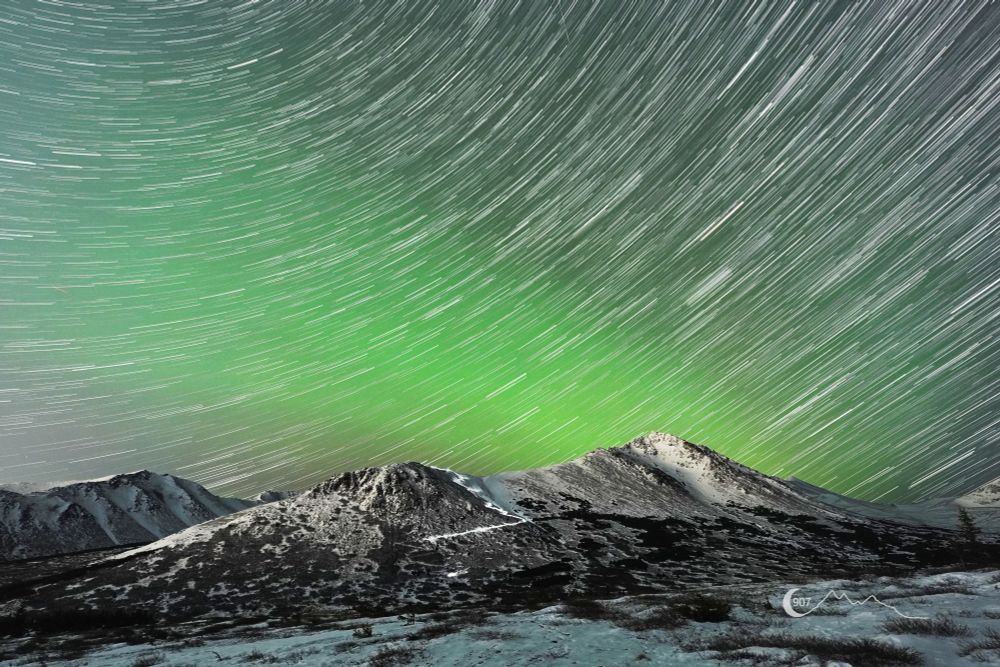 Star trails arc over the Chugach Mountains, with a faint green aurora band glowing above the mountains. A single light trail from a hiker's headlamp winds its way up the mountainside.