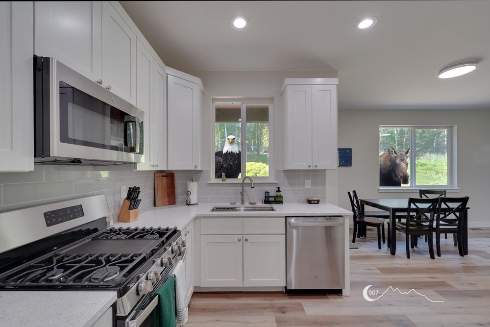 A photograph of a kitchen looking towards a window above a kitchen sink and a window above the dining room table. An eagle is looking in the window above the kitchen sink and a moose is looking in the window above the kitchen table.