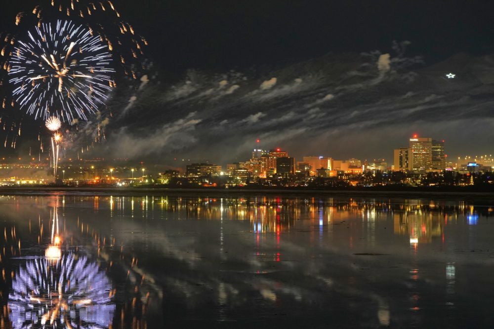 A photograph of  fireworks near downtown Anchorage. The fireworks are glowing orange and blue to the left of the main downtown areas. Buildings are glowing in the dark as smoke from the fireworks drifts over the tops of the buildings. The scene reflects perfectly in the water surrounding downtown.