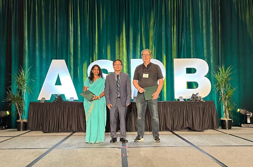 Three people stand side-by-side on a stage with large letters behind them, "ASPB." Two people hold certificates in their hands.