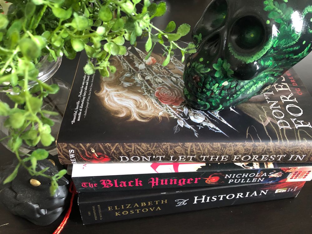 A stack of books (Don’t Let the Forest in by CG Drews, a The Black Hunger by Nicholas Pullen, and The Historian by Elizabeth Kostova) sit next to a fake fern plant. On top of the stack is a skull painted with vines and leaves with glowing green eyes. 