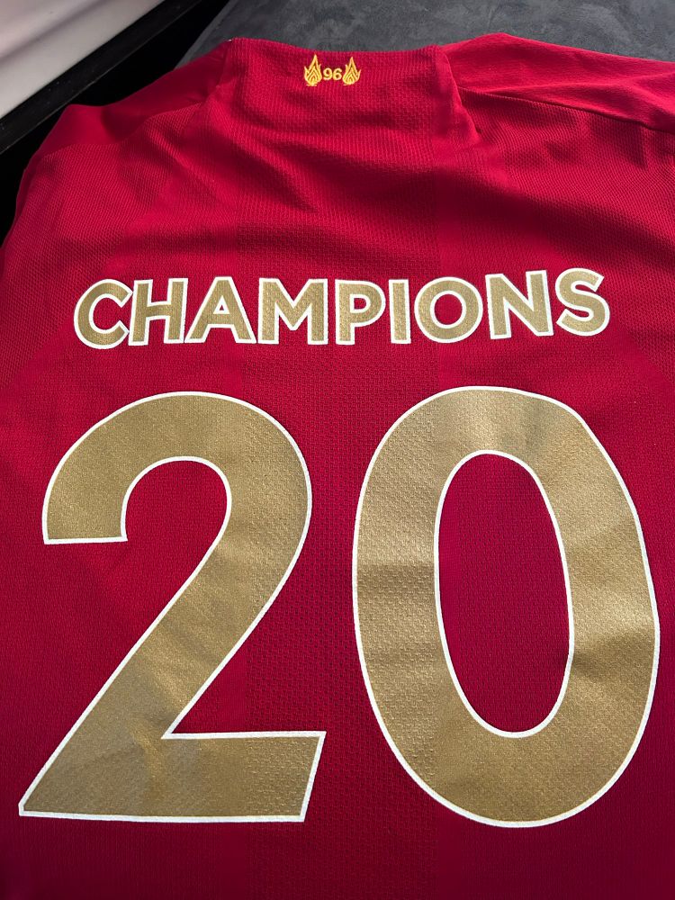 The back of a LFC red home jersey with CHAMPIONS 20 in gold lettering. It also has the eternal flames and the number 96 below the collar to remember the (then) 96 fans who’d died at Hillsborough. 