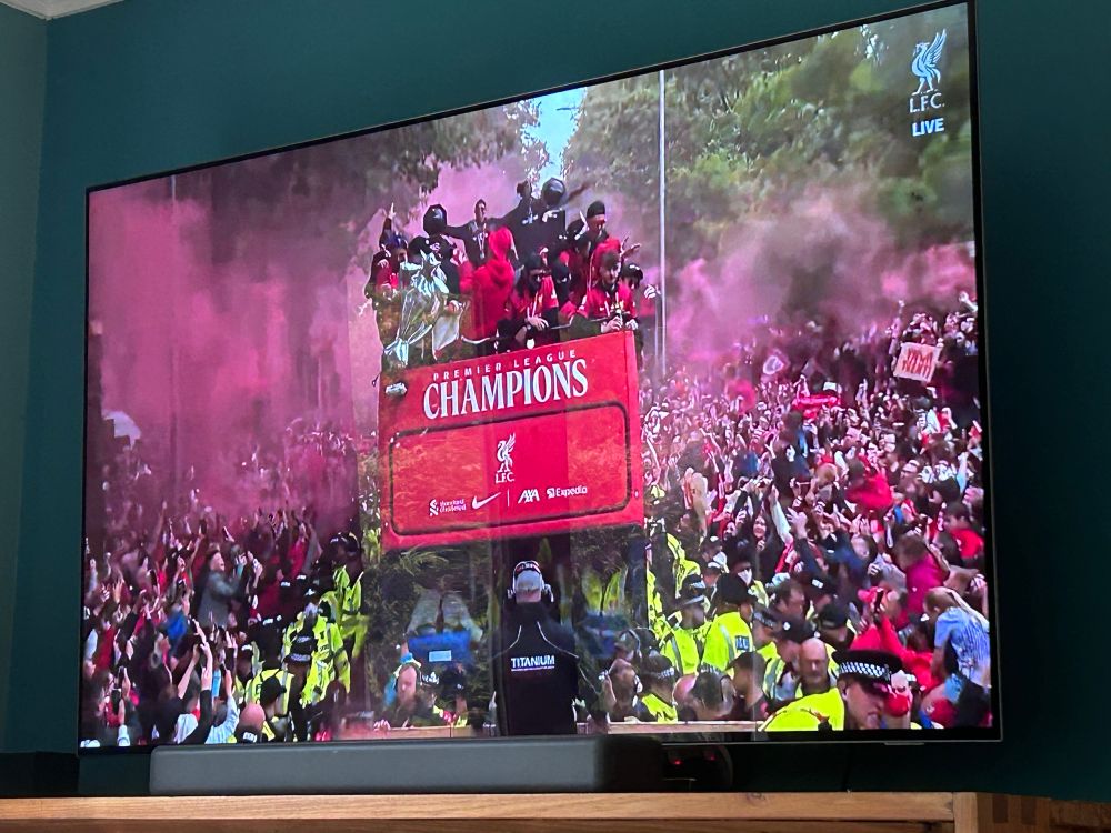 Liverpool men’s team on board a red bus that says Premier League champions as it makes its way through a crowd of fans. There is red smoke in the air and the bus is escorted by police as it makes its way through the city. 