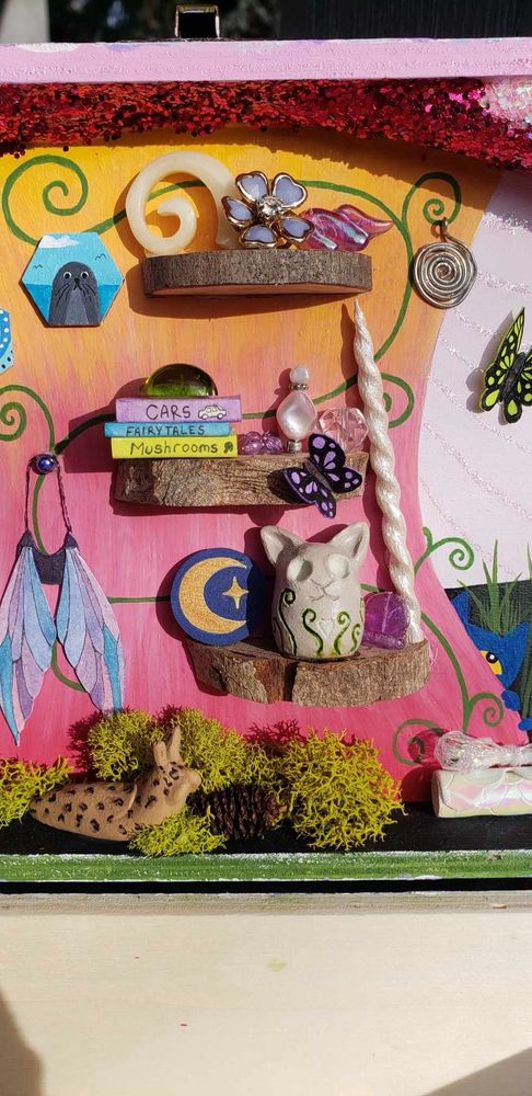 Close up of the middle of the lid diorama fairy house including wooden shelves made from thin slices of a tree branch, hand made and painted clay cat statue and leopard slug, paper fairy wings, hand made wooden and painted unicorn horn, hand made books and some little jewels. Also a painting of a Selkie and moon and stars