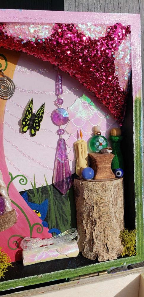 Close up of the right side of the fairy house diorama in the lid of a trinket or treasure box. Includes Pete the cat hiding behind the trunk of the mushroom, iridescent mermaid or dragon scales, a tree stump with potion bottles a candle and some beads on it, little bits of moss tucked in around the tree trunk and a jewel hanging from the roof of the mushroom/fairy house. Also a paper butterfly.
Everything hand made!