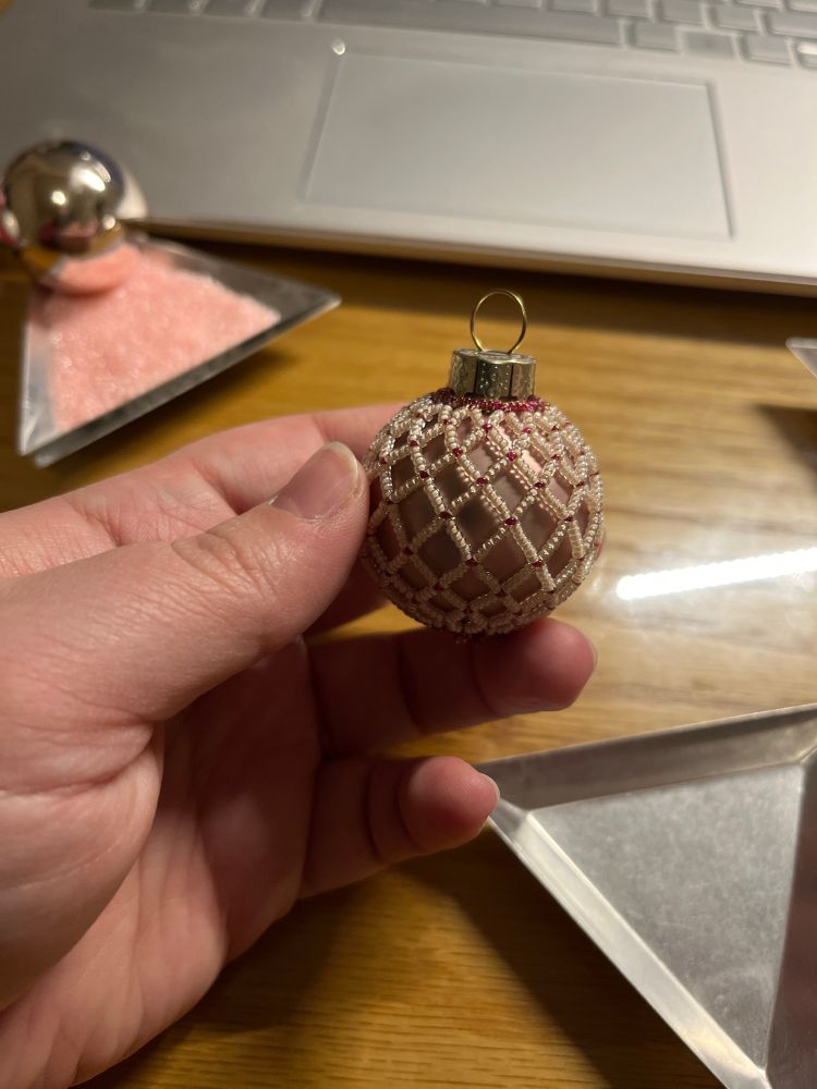 A hand rests on a desk near a palm-sized triangular metal dish and a laptop keyboard. The hand holds a spherical pale taupe glass ornament, covered in an open netting of tiny seed beads in a matching pale off-white with cranberry-red accents. 