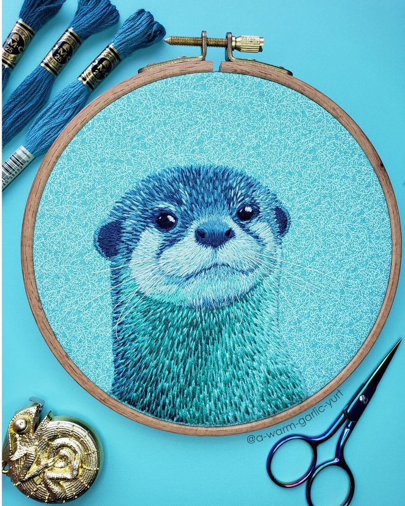 Hand embroidery of an otter, stitched in greens and blues, in a hoop surrounded by floss, scissors, and measuring tape