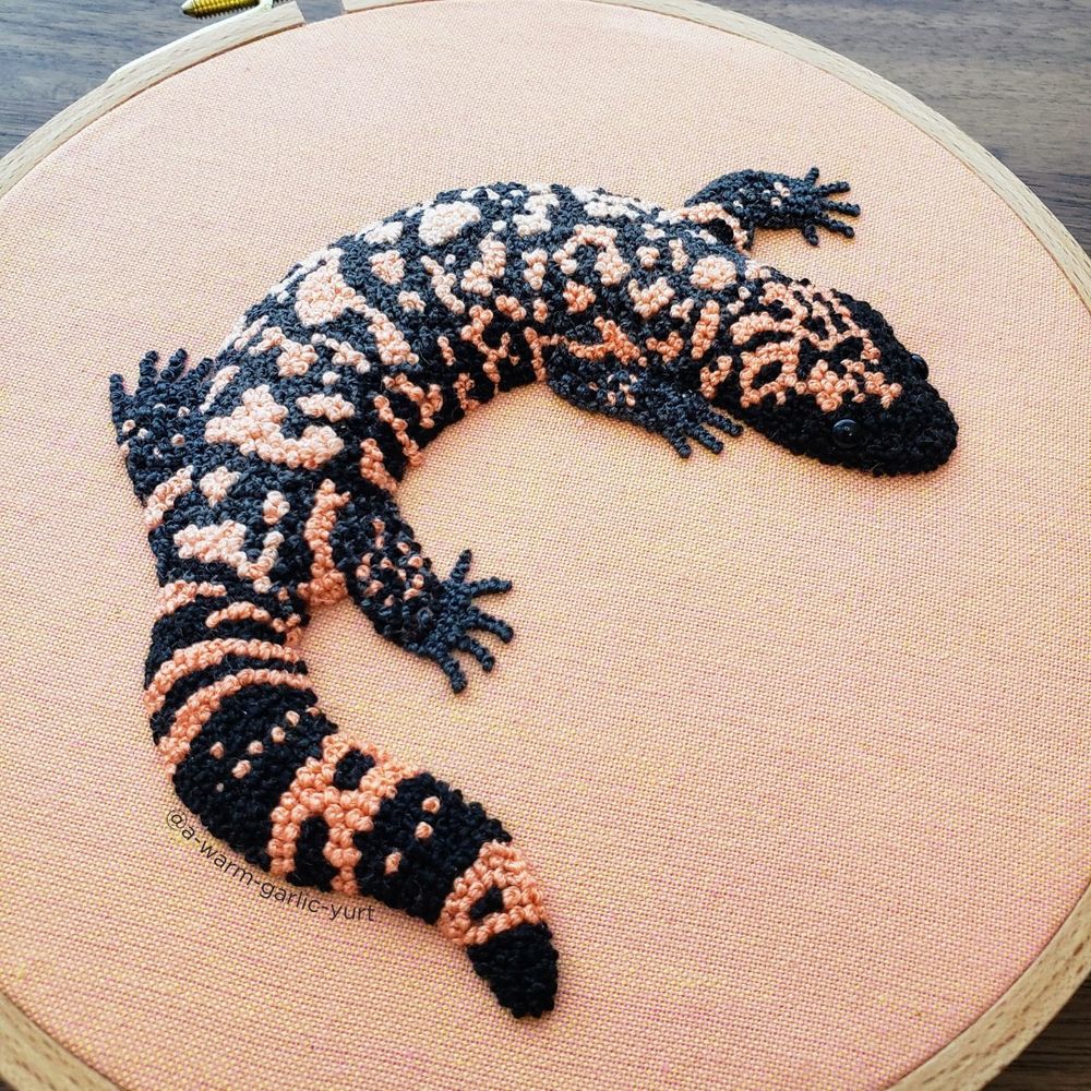 Hand embroidery of a gila monster, stitched with thousands of French knots in salmon and black on salmon fabric, framed in a wooden hoop. The stitching is raised on the fabric giving it a 3D effect