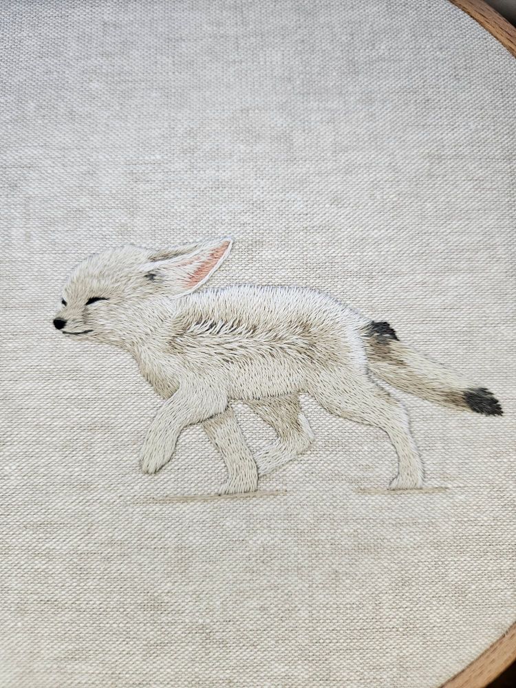Hand embroidery of a fennec fox, stitched in light neutral tones, walking in a strong wind