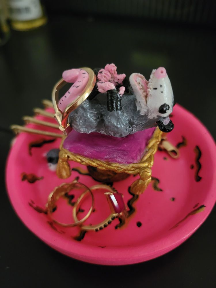 Taken from the front, a hot pink dish with black and gold squiggles, a small sculpture of an opossum on their back with their tail curled up to touch their feet. Mouth is joyfully open. They lay on a fuschia pillow with gold trim and tassels slightly above the bottom of the dish, raised up a bit by a black ball with gold brushed on top. Inside the dish there is a tiny opossum. There's a gold ring hanging from the opossum tail, two more rings in the dish and then four gold hair pins with pearls hanging over the side of the dish. 