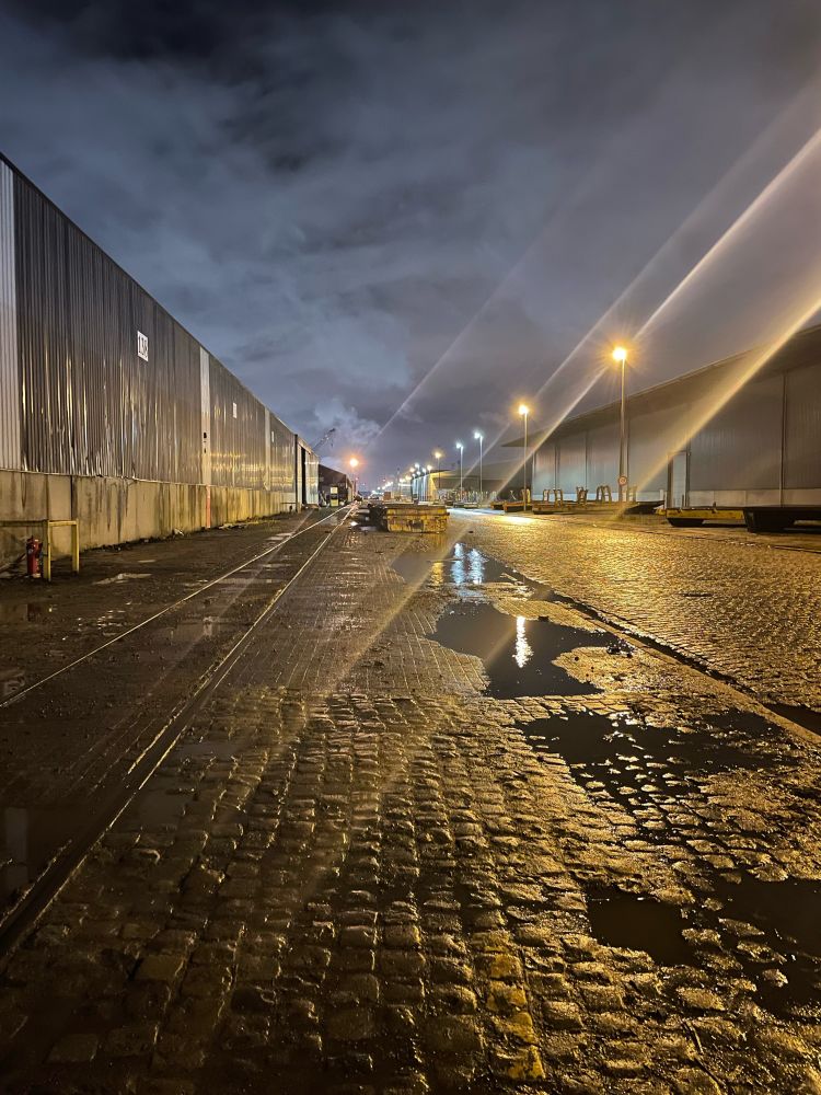 A cobble stone street, with puddles of water, street lights and warehouses along both sides. No one around at this dock in Antwerp Harbour at night. A cloudy sky, lit by artificial light. Eerie atmosphere. 